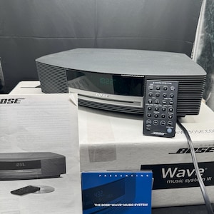 Puede incluir: Un sistema de música Bose Wave negro con mando a distancia, manual y caja. El sistema muestra la hora 10:22. La caja y el manual tienen el logotipo de Bose e información del producto. El mando a distancia está etiquetado como "3508".