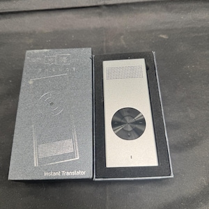 Puede incluir: Un traductor instantáneo plateado con un botón de control negro se muestra junto a su caja gris. La caja tiene las palabras "Instant Translator" impresas. El dispositivo es rectangular con una rejilla de altavoz en la parte superior.