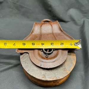 May include: A rusty metal pulley with a wooden wheel, measuring 4 1/2 inches in diameter. The pulley has a metal hook and a metal pin for attaching a rope.