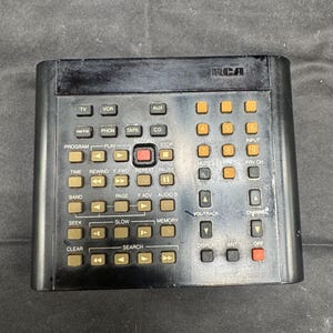 Op de afbeelding: Een zwarte RCA universele afstandsbediening met talrijke knoppen voor tv, VCR, cd en andere functies. De afstandsbediening heeft oranje en witte tekstlabels voor diverse bedieningselementen.