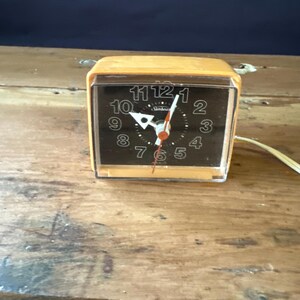 May include: A vintage orange alarm clock with a black face and white numbers. The clock has a clear plastic cover and a cord.