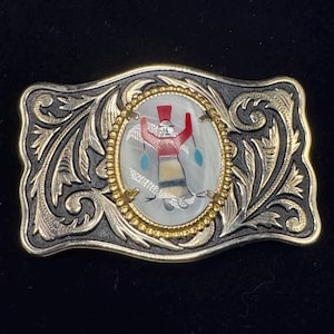 May include: Rectangular silver-tone belt buckle with an oval mother-of-pearl inlay featuring a Native American figure in red and turquoise. The buckle has a gold-tone border and ornate scrollwork.