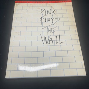 Könnte beinhalten: Ein weißes Buch mit einem Ziegelmuster auf dem Cover. Der Titel "Pink Floyd The Wall" ist mit schwarzem Stift auf dem Cover geschrieben. Das Buch ist eine Gitarrentabulaturedition.