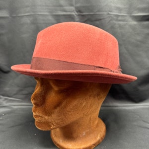 Pode incluir: Um chapéu fedora bordô com uma faixa de fita preta. O chapéu está em uma cabeça de manequim marrom.