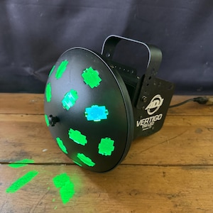 May include: Black DJ lighting fixture with green and blue lights. The fixture has a circular front with green square-shaped lights and the text "VERTIGO TRI LED". The fixture is on a wooden surface.