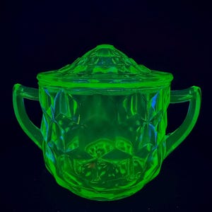 Puede incluir: Un azucarero de vidrio verde brillante con tapa y dos asas. El cuenco y la tapa tienen un patrón de diamantes. El azucarero está sobre un fondo oscuro.