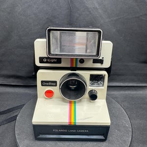 May include: Cream-colored Polaroid OneStep Land Camera with a black base and flash. Features a rainbow stripe and the text "Polaroid Land Camera." The flash unit is labeled "Q-Light."