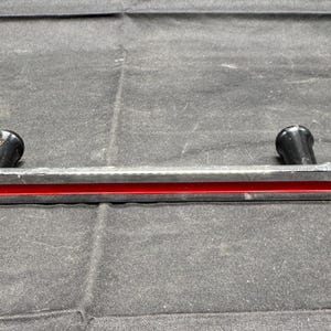 May include: A silver-colored metal handle with a red stripe and two black knobs. The handle has a curved design and is set against a dark gray background.