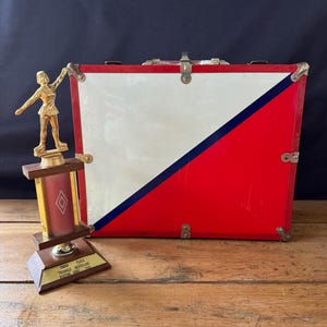 May include: A gold-colored trophy with a figure skater on top. The trophy has a wooden base with a red and gold plaque that reads "Third Place Triangle Skateland Dayton, Ohio 1968". A red, white, and blue metal box is in the background.