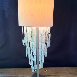 May include: A decorative table lamp with a beige lampshade and a silver-colored base. The lamp features a cascade of rectangular, iridescent glass pieces hanging from the shade, creating a unique lighting effect. The base is square.