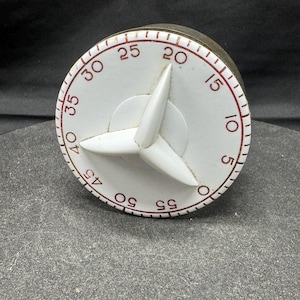 May include: A vintage white kitchen timer with a red-numbered dial and a three-winged white knob. The timer's face displays numbers from 5 to 55, with a dark brown base. The timer is designed to sit on a flat surface.