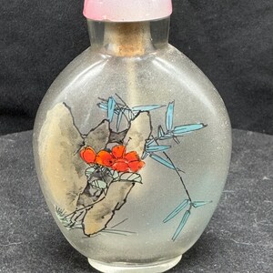 May include: A clear glass perfume bottle with a pink stopper. The bottle features a hand-painted design of a red flower, bamboo, and a rock formation. The bottle has a cork stopper and a rounded shape.