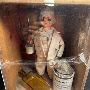 Pode incluir: Uma boneca vintage de um pintor numa caixa de madeira. A boneca usa um uniforme bege, óculos e um boné. Uma lata de tinta, uma garrafa e um raspador de tinta também estão na caixa. A lata de tinta diz "Beatty Paints COOL-KRETE RED LATEX".