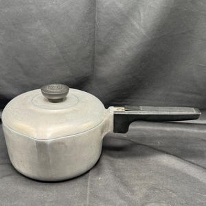 May include: A vintage aluminum saucepan with a lid and a black handle. The pot has a rounded body and a lid with a black knob. The handle is attached to the side of the pot. The saucepan is likely used for cooking.