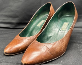 May include: A pair of brown leather heeled shoes with a pointed toe. The shoes have a green leather lining and the word "Gucci" is printed on the inside of the shoes.