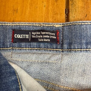 May include: Close-up of a blue denim jean label with white stitching. The label reads "High Rise Tapered Skinny Tres &Eacute;troite Jambe Effil&eacute;e Taille Haute" and "COLETTE" in red.