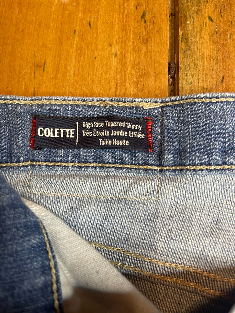 May include: Close-up of a blue denim jean label with white stitching. The label reads "High Rise Tapered Skinny Tres &Eacute;troite Jambe Effil&eacute;e Taille Haute" and "COLETTE" in red.