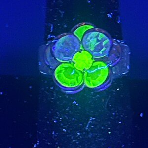 May include: A flower-shaped ring with five green stones set in a silver band. The stones are glowing under a black light.
