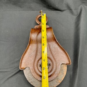 May include: A rusty metal pulley with a wooden wheel, measuring approximately 11 inches in diameter. The pulley has a metal hook at the top and a metal ring around the center.