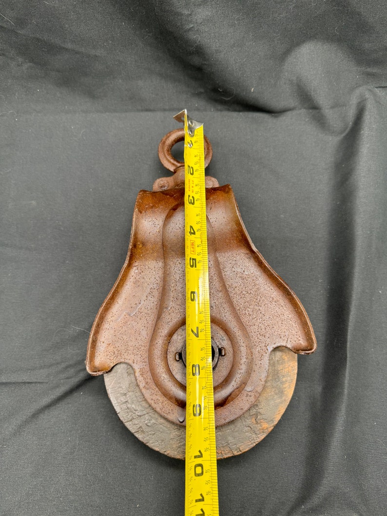 May include: A rusty metal pulley with a wooden wheel, measuring approximately 11 inches in diameter. The pulley has a metal hook at the top and a metal ring around the center.