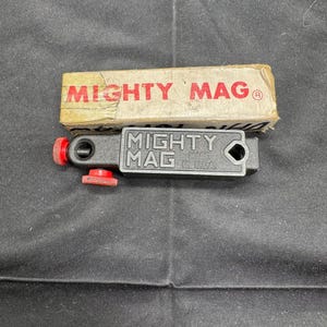 Puede incluir: Una herramienta Mighty Mag negra y roja con una perilla de ajuste roja y el texto "MIGHTY MAG MADE IN U.S.A." se muestra junto a una caja de cartón con el texto "MIGHTY MAG" en rojo.
