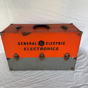 May include: Vintage orange and gray General Electric Electronics toolbox. The box has a black handle, metal latches, and corner protectors. The words "GENERAL ELECTRIC ELECTRONICS" are printed in black on the front.