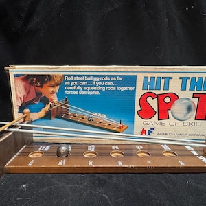 May include: A vintage Hit The Spot game of skill featuring a wooden base with circular holes, two long metal rods, and a steel ball. The original cardboard box displays the game title, the American Toy & Furniture Company logo, and instructions for rolling the ball uphill by squeezing the rods.