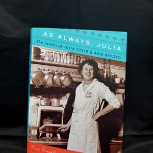 Peut inclure: Un livre à couverture rigide intitulé "AS ALWAYS, JULIA" avec une couverture turquoise et rouge. La couverture présente une photo en noir et blanc de Julia Child dans une cuisine. Le sous-titre du livre est "The Letters of JULIA CHILD & AVIS DEVOTO".