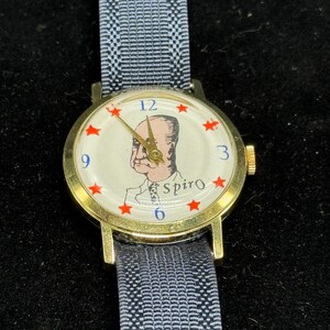 May include: A vintage gold-tone wristwatch with a light-colored face featuring a portrait and the name "Spiro." Red star hour markers are present. The watch has a dark gray and white patterned band.