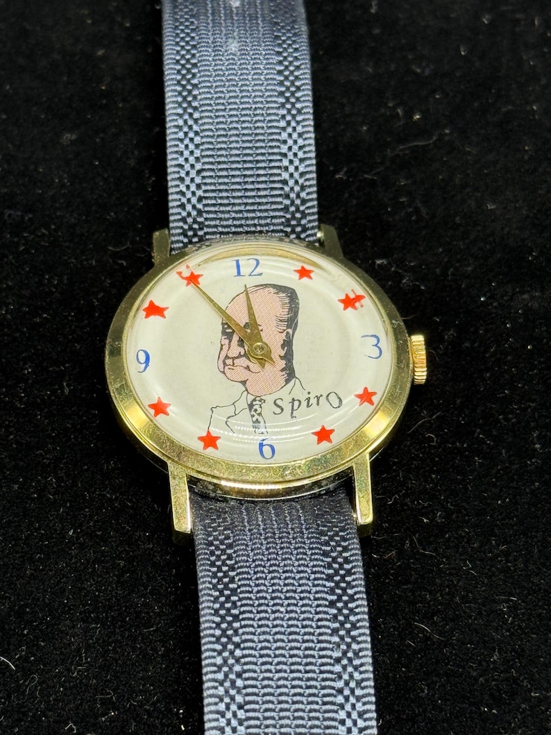 May include: A vintage gold-tone wristwatch with a light-colored face featuring a portrait and the name "Spiro." Red star hour markers are present. The watch has a dark gray and white patterned band.