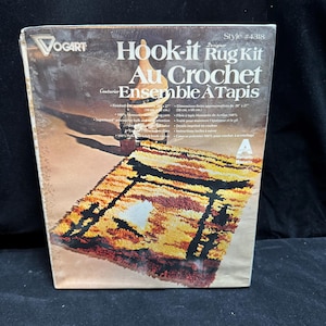 May include: A Vogart Hook-it Rug Kit, style #4318, featuring a finished rug design in shades of yellow, orange, and black. The packaging displays the text Hook-it Rug Kit, Au Crochet Ensemble A Tapis, and lists finished dimensions of 20 x 27 inches.