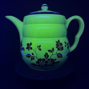Può includere: Una teiera verde brillante con coperchio e manico. La teiera presenta un motivo floreale marrone scuro. La teiera è appoggiata su una superficie scura. Si tratta di un oggetto vintage.