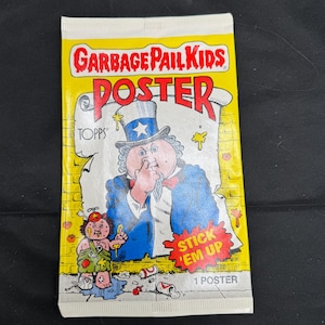 Può includere: Un pacchetto di poster Garbage Pail Kids con sfondo giallo e le parole "GARBAGE PAIL KIDS POSTER" in rosso e bianco. Il pacchetto presenta un'illustrazione di Zio Sam e un personaggio Garbage Pail Kid. È visibile anche il testo "STICK 'EM UP".