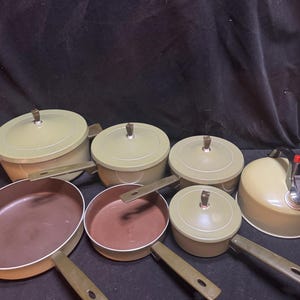 May include: A set of kitchen cookware including two large pots with lids, two smaller pots with lids, one large frying pan, one small frying pan, and a tea kettle. The cookware features a light olive green exterior, brown interior surfaces, and brown handles.