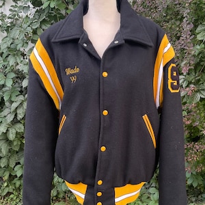 May include: Black varsity jacket with yellow and white stripes on the sleeves. The jacket has a gold button closure and a pocket on the left side. The text "Wade '99" is embroidered on the left chest and the number "6" is embroidered on the right sleeve.