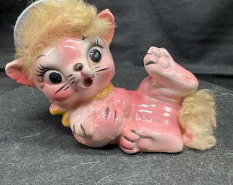 May include: A vintage pink ceramic cat figurine with a surprised expression. The cat has large black eyes, a yellow collar, and a tuft of blonde fur on its head and tail. The figurine is lying on its back.