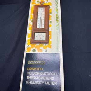 Puede incluir: Un termómetro interior-exterior y un higrómetro vintage enmarcados en madera con una esfera blanca y números negros. El termómetro tiene un indicador rojo y azul. El higrómetro tiene una aguja negra. El marco es marrón con un ribete dorado. La caja es blanca con texto negro que dice "Springfield Lynwood Termómetros interior-exterior e higrómetro".