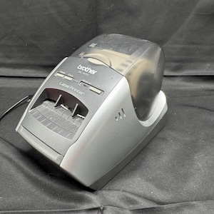 Puede incluir: Una impresora de etiquetas Brother QL-570 plateada y negra. La impresora de etiquetas tiene una cubierta transparente y una ranura para etiquetas. El texto "Label Printer" es visible en la parte frontal.