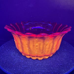 May include: A round glass bowl with a scalloped, petal-like rim, glowing with a vibrant orange and red hue under ultraviolet light. The exterior features a vertical ribbed texture, while the interior is smooth and deep, reflecting the light.