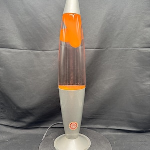 Könnte beinhalten: Retro-Lavalampe mit silberfarbenem Sockel und Oberteil sowie einem transparenten Glaskörper. Die Lampe enthält orangefarbene Lava. Die Lampe ist eingesteckt und einsatzbereit. Die Lampe ist ca. 40 cm hoch.