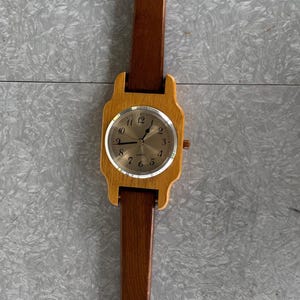 May include: Wooden wristwatch with a rectangular face and a brown leather strap. The watch face has a light-colored background with black numerals and hands. The watch band is made of dark brown wood.