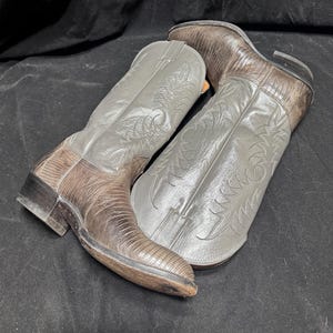 Könnte beinhalten: Ein Paar zweifarbige Cowboystiefel mit einem dunkelbraunen, strukturierten Unterteil und einem glatten grauen Lederschaft. Die grauen Schäfte sind mit aufwendigen, tonalen Stickmustern verziert und die Stiefel haben einen niedrigen Absatz.
