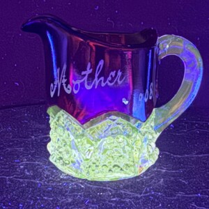 May include: A glass creamer pitcher featuring a vibrant yellow-green glow under ultraviolet light. The upper portion is dark with the word Mother etched in script, while the lower section displays a textured, diamond-patterned surface.