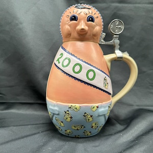 Peut inclure: Une chope à bière en céramique en forme de personne portant un motif de couche-culotte bleu et blanc. La chope a un corps brun clair avec une écharpe bleu et blanc qui dit "2000". La chope a un couvercle avec un cadran d'horloge et une poignée.