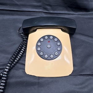 May include: A vintage beige and black telephone with a rotary dial. The phone has a black handset and a coiled cord. The dial features black numbers and letters, with a red light above the HOLD button. A classic communication device.