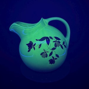 Puede incluir: Una jarra de cerámica verde claro vintage con un cuerpo redondeado y un asa curva. La jarra presenta un diseño floral en azul oscuro y gris. La jarra tiene un pico para verter líquidos. El fondo es azul oscuro.