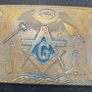 May include: Rectangular brass belt buckle with Masonic symbols. The buckle features a central square and compass design with a blue "G", surrounded by other symbols like an all-seeing eye, hammer, and trowel. The buckle has a worn, antique appearance.