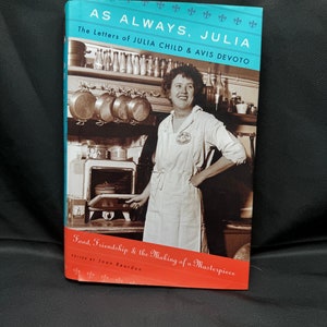 Peut inclure: Un livre intitulé "As Always, Julia: The Letters of Julia Child & Avis Devoto" avec une couverture rouge et une photo de Julia Child sur la couverture. Le livre parle de la nourriture, de l'amitié et de la création d'un chef-d'œuvre.
