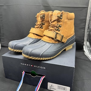May include: Tan leather boots with a blue rubber bottom and a buckle strap. The boots are sitting on a black box with the Tommy Hilfiger logo and the words "Medium Blue, 5Y" and "The Reginald 8 M" printed on the box.
