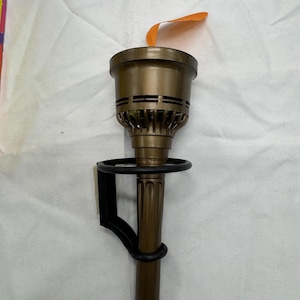 May include: A bronze-colored torch with a black bracket. The torch has a cylindrical handle and a flared top with horizontal lines. An orange flame-shaped piece is attached to the top. The torch is designed to be mounted on a wall.